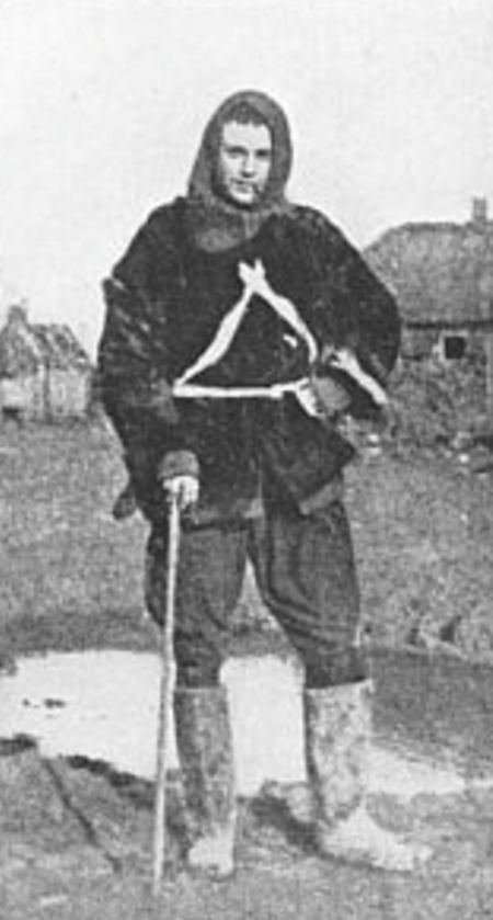 CAPTAIN BRUCE BAIRNSFATHER This picture was taken at the Front, less than a quarter of a mile from the German trenches. Captain Bairnsfather has come "straight off the mud," and is wearing a fur coat, a Balaclava helmet, and gum boots. Immediately behind him is a hole made by a "Jack Johnson" shell.