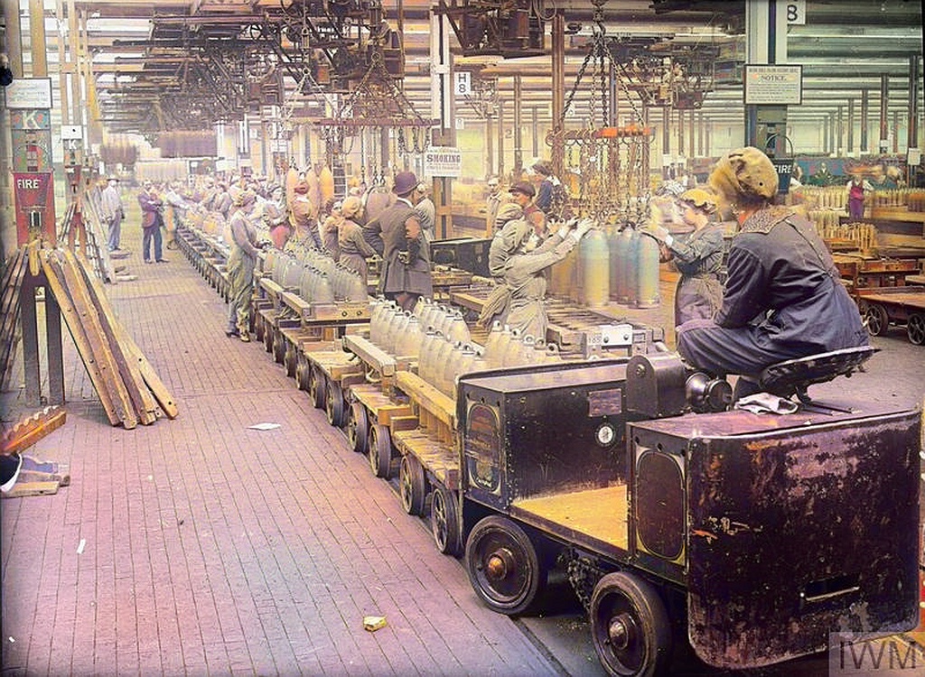 A Woman Drives A Trolley Train Across A Busy Factory Floor At The National Filling Factory, Chilwell. The Trolley Is Loaded With Shells And Is Used To Transport The Shells From One Part Of The Factory